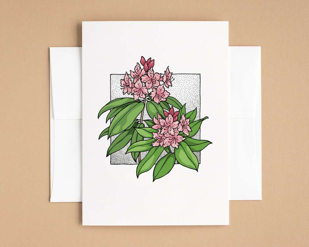 A vertical white card depicts two pink rhododendron flower clusters with green leaves. The card sits on top of a white envelope, which lies on top of a brown backdrop.